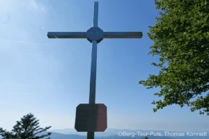Gipfelkreuz auf einem Berg mit Blick auf die Berge im Hintergrund.