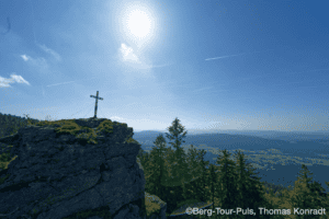 Berggipfel mit Kreuz, Blick auf das Tal und die Berge bei Sonnenschein.