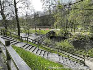 Naturpfad mit Wasser, Brücke und Treppen im Grünen.