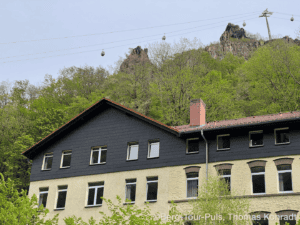 Historisches Haus vor grüner Bergkulisse mit Seilbahn und Felsen.
